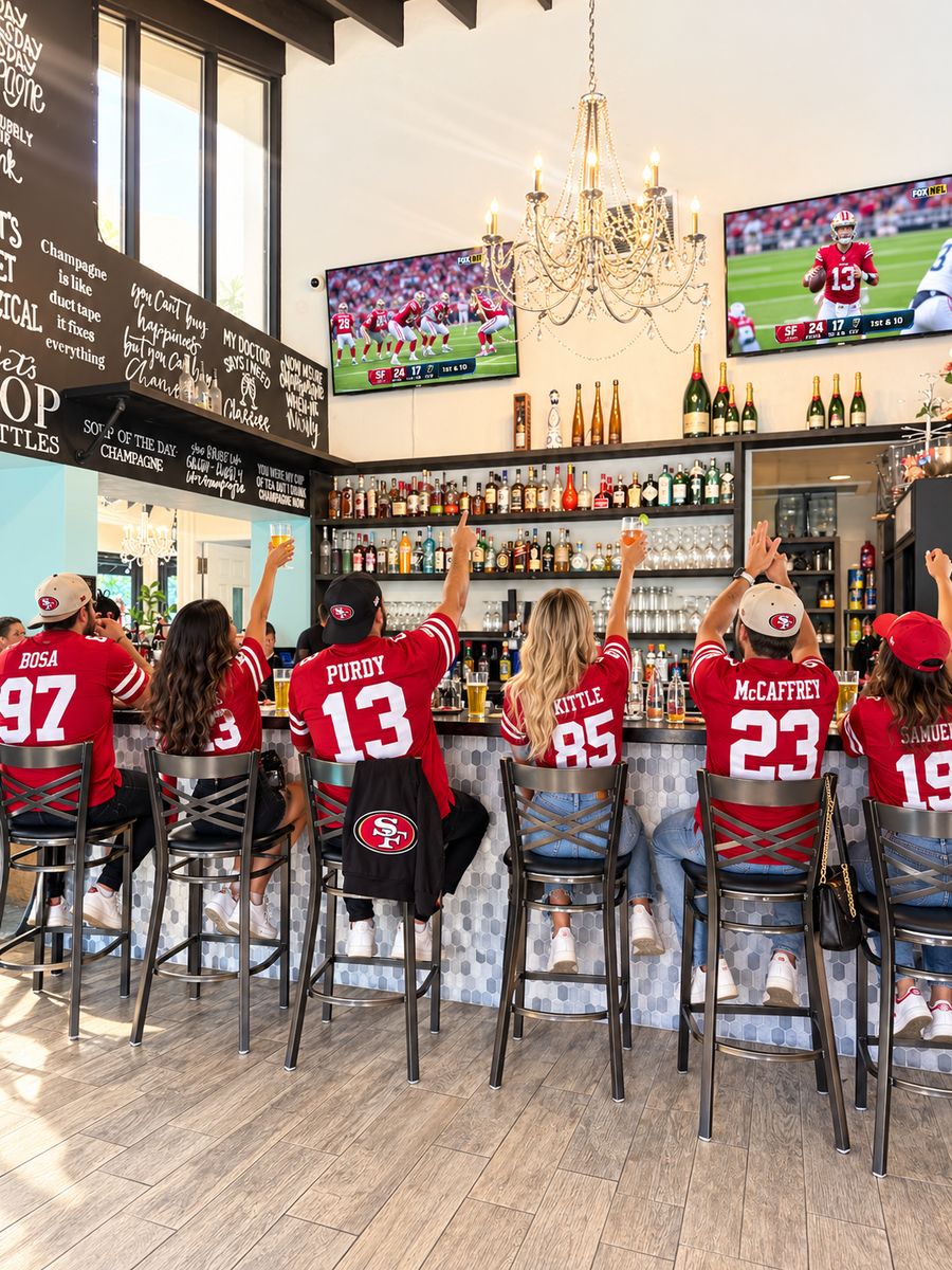 Friends in red football jerseys raising their glasses at a Mimosa House bar under a crystal chandelier, the game on the TVs above